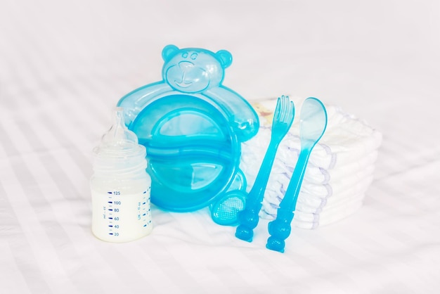 Set da tavola per bambini latte biberon nibbler posate blu cucchiaio forchetta pannolini pannolini usa e getta sul letto bianco sfondo vista frontale