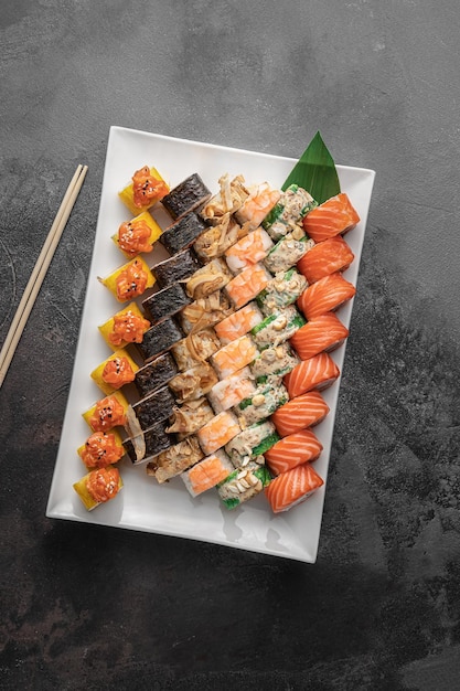 Set di diversi involtini di sushi maki colorati con tonno salmone gamberetti granchio e avocado e foglia di bambù verde in un piatto di ceramica bianca con bacchette su uno sfondo grigio scuro testurizzato vista dall'alto