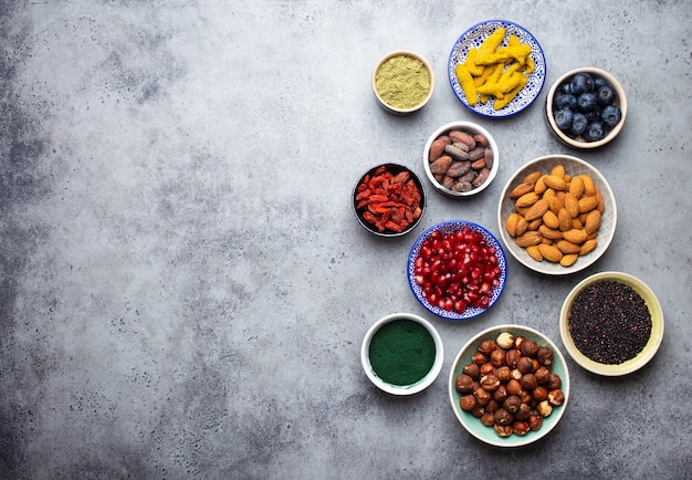 Set di diversi supercibi in ciotole su sfondo grigio pietra: spirulina, bacche di goji, cacao, tè verde matcha, quinoa, semi di chia, mirtilli, noci per una vita felice e sana, vista dall'alto, spazio copia