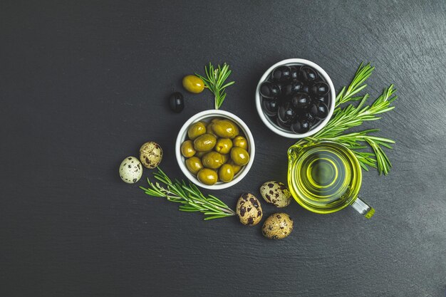 Set di olive nere e verdi uova di quaglia su piatti olio d'oliva e rosmarino su sfondo di pietra nera Spazio per la copia vista dall'alto