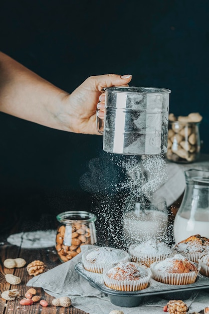 Setacciare lo zucchero a velo su un delizioso cupcake con uvetta, mandorle e noci in cucina Mano femminile invecchiata che setaccia lo zucchero a velo con un setaccio sopra muffin al caramello alla vaniglia in bicchieri di carta in una teglia