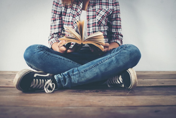 Foto sezione bassa di una giovane donna che tiene un libro mentre è seduta contro il muro sul pavimento di legno