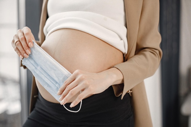 Sezione mediana della donna incinta con maschera facciale in piedi vicino alla finestra in ufficio