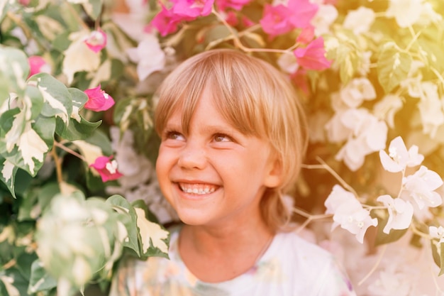 Sfocato ritratto sfocato del viso candido poco felice sorridente bambino biondo di cinque anni ragazzo con gli occhi verdi in piante da fiore rosa e bianco in natura i bambini si divertono il bagliore delle vacanze estive