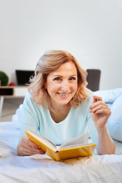 Sfogliando le pagine. Allegra donna dai capelli chiari che riposa sul suo letto e legge un libro interessante mentre è sola a casa