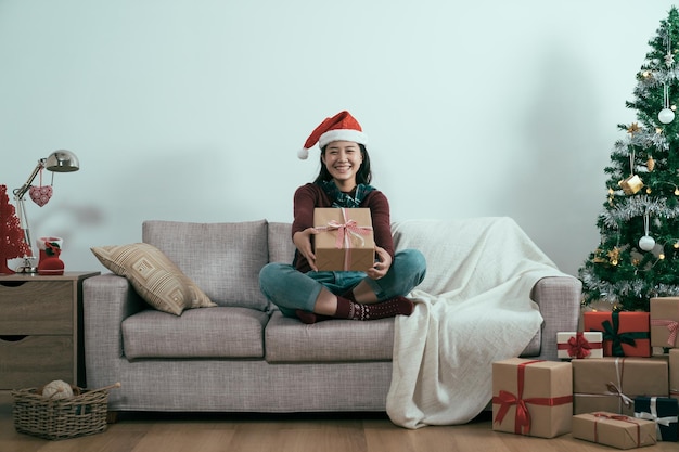 sfondo bianco all'interno. felice giovane donna giapponese asiatica in cappello rosso santa dando un regalo di Natale a casa soggiorno divano faccia fotocamera. affascinante ragazza indossare maglione sorridente inviando presente nel divano