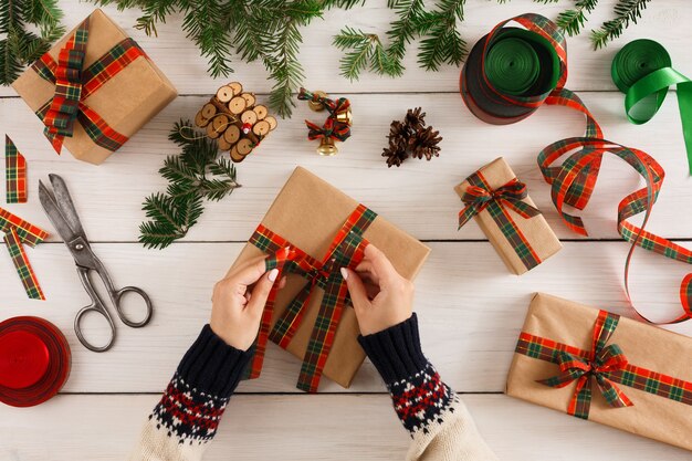 Sfondo di confezioni regalo. Vista dall'alto sulle mani femminili confezione regalo di Natale in carta artigianale, decorare con nastro di raso al tavolo di legno rustico disordinato bianco. Concetto di vacanze invernali.