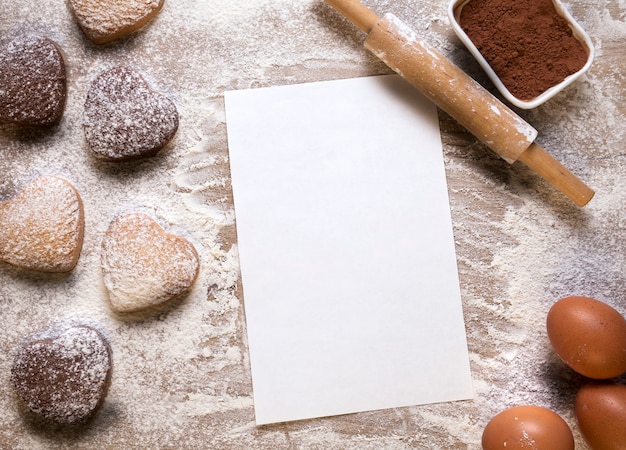 Sfondo di cottura con foglio di carta bianco per la ricetta o il menu, biscotti a forma di cuore, uova, farina e mattarello. Spazio vuoto per il testo. San Valentino