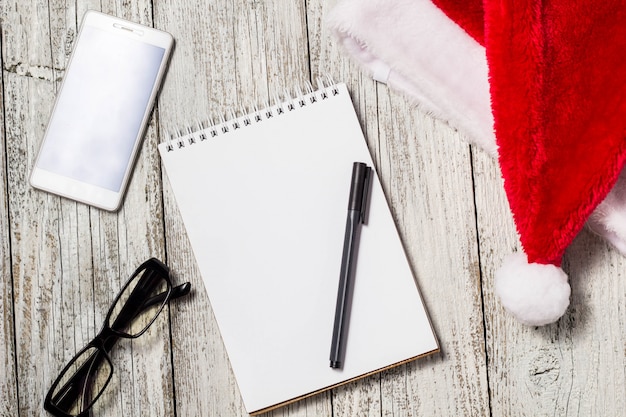 Sfondo di Natale e Capodanno con smartphone, cappello rosso di Babbo Natale, occhiali, blocco note e penna per i tuoi desideri