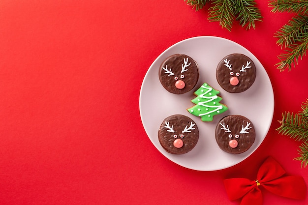 Sfondo di Natale rosso con biscotti al cioccolato decorati con facce di cervo e abete, vista dall'alto, layout piatto