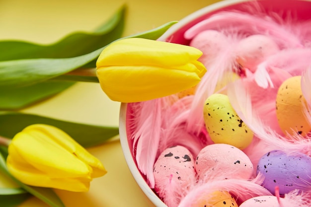 Sfondo di Pasqua festivo Ciotola rosa con uova colorate piume rosa e tulipani gialli Buon concetto di Pasqua Cartolina postale su sfondo giallo