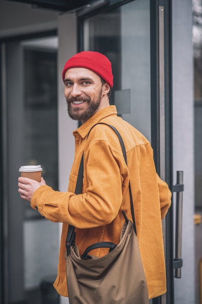 Sguardo allegro. Uomo con un cappello rosso e una giacca arancione con una tazza di caffè in mano che sembra allegro