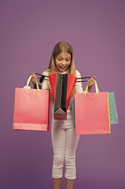 Sguardo del bambino in borse della spesa su sfondo viola Piccolo sorriso da maniaco dello shopping sorpreso con sacchetti di carta Infanzia Shopping e vendita Shopper ragazza con sacchetti di carta Che sorpresa Grandi saldi e shopping