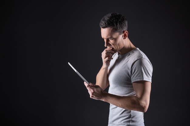 Sguardo premuroso. Uomo premuroso piacevole intelligente in piedi con un tablet in mano e tenendo il mento mentre pensa al lavoro