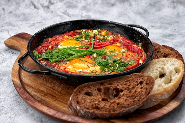 Shakshuka su una padella e una tavola di legno, con cipolle verdi, peperoni e crostini di pane