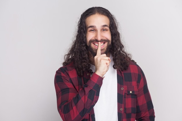 Shh, questo è segreto! uomo divertente con barba e capelli neri lunghi ricci in camicia rossa a scacchi casual in piedi e mostrando segno silenet per segreto a sorpresa. foto in studio al coperto, isolata su sfondo grigio