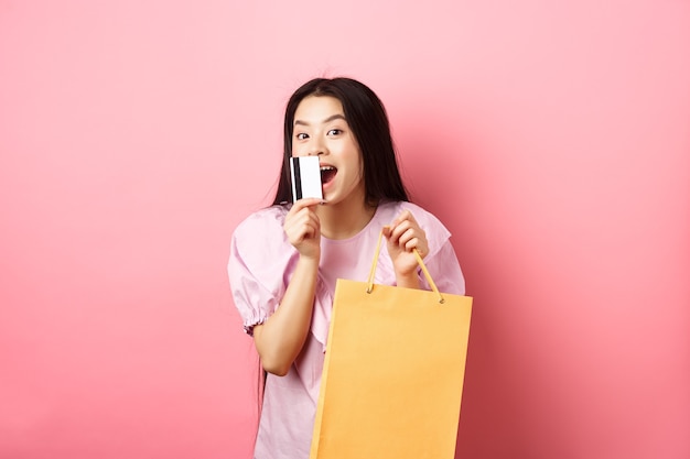 Shopping. Bella donna asiatica emozionante che compra nei negozi, che tiene il sacchetto di carta e che bacia la carta di credito di plastica, levantesi in piedi contro il fondo rosa.