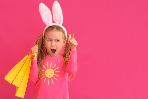 Shopping di Pasqua Eccitato emozione sorpresa adorabile bambina in costume da coniglio orecchie da coniglio tenere borse della spesa gialle isolato sfondo rosa con il dito puntato su Kid Sale sconti pasqua bambini