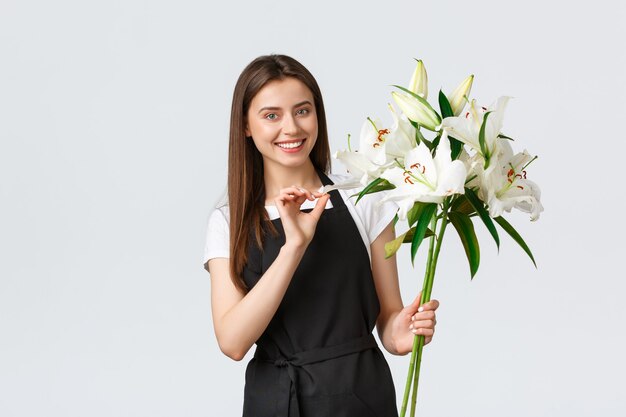 Shopping, dipendenti e concetto di piccola impresa. Sorridente bella fiorista commessa in un negozio di fiori che tiene in mano un mazzo di gigli bianchi e sorride, prepara l'ordine per il cliente