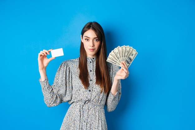 Shopping. Elegante donna impertinente con banconote da un dollaro e carta di credito in plastica, guardando fiducioso alla macchina fotografica, blu.