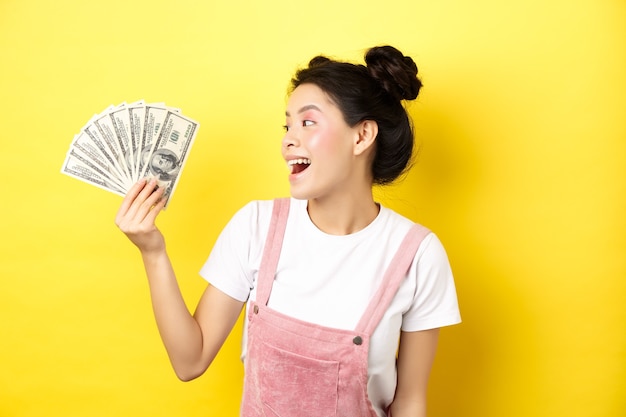 Shopping. Modello femminile asiatico ricco ed elegante che mostra soldi, guardando le banconote da un dollaro con la faccia felice, in piedi sul giallo