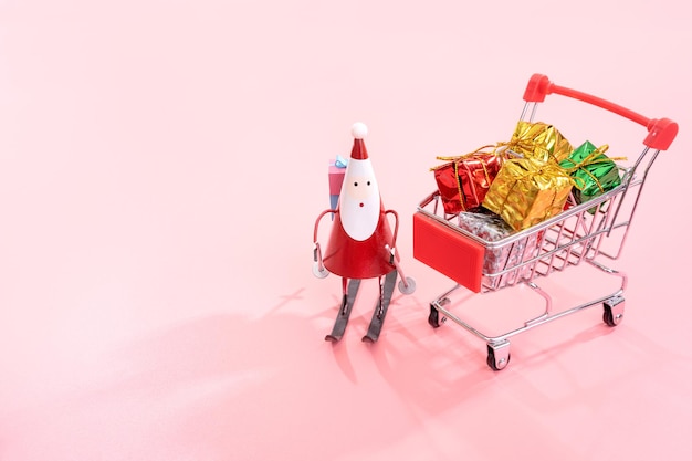 Shopping natalizio concetto mini carrello rosso carrello con Babbo Natale giocattolo e confezione regalo isolato su sfondo rosa pallido spazio vuoto per la copia da vicino