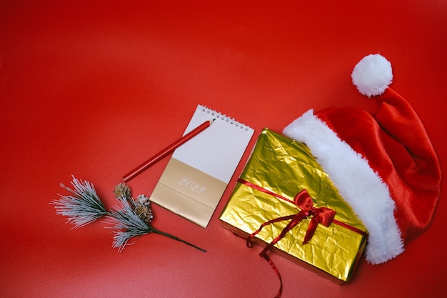 Shopping natalizio. Elenco degli acquisti e dei regali per il nuovo anno. Il cappello di Babbo Natale giace su una scatola d'oro su sfondo rosso.