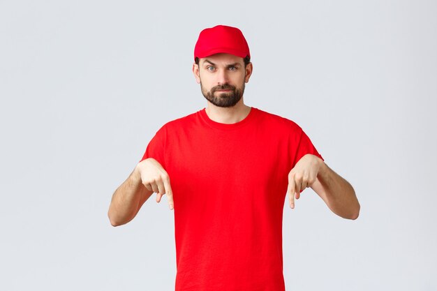 Shopping online, consegna durante la quarantena e concetto da asporto. Corriere scettico o sospettoso con berretto e t-shirt uniforme rossa, con le dita puntate verso il basso, sfondo grigio in piedi