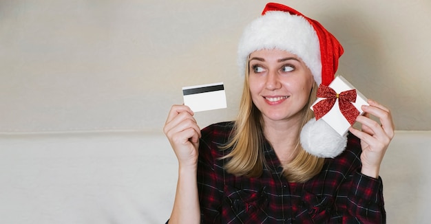 Shopping online di Natale. Femmina che tiene una carta e una confezione regalo. Donna che indossa un cappello rosso di Natale, acquista regali, si prepara al Natale.