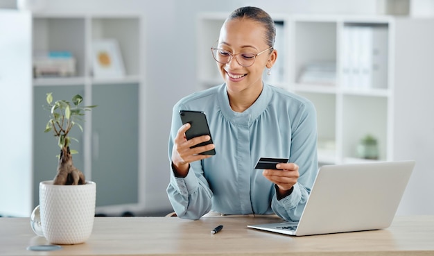 Shopping online esecutivo femminile che acquista con una carta di credito aziendale su un telefono Una donna sorridente che paga le bollette aziendali seduto alla scrivania dell'ufficio con il laptop Signora felice che effettua un acquisto su un sito Web