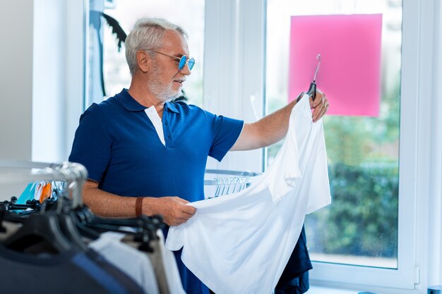 Shopping. Profilo di elegante uomo dai capelli grigi positivo in piedi nel negozio mentre si sceglie una nuova maglietta bianca con curiosità
