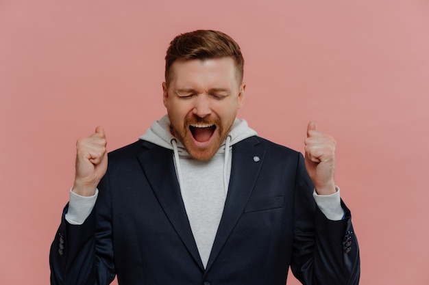 Sì, l'ho fatto. Studio shot di un uomo rosso felice felicissimo in abbigliamento casual che tiene gli occhi chiusi dalla felicità e alza i pugni chiusi mentre celebra il successo, le persone che raggiungono obiettivi personali