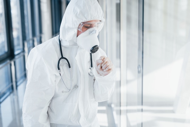 Si sente male e male. Scienziato medico femminile in camice da laboratorio, occhiali difensivi e maschera in piedi al chiuso