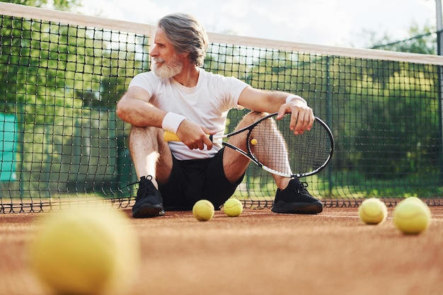 Si siede a terra e si prende una pausa Uomo anziano moderno ed elegante con racchetta all'aperto sul campo da tennis durante il giorno