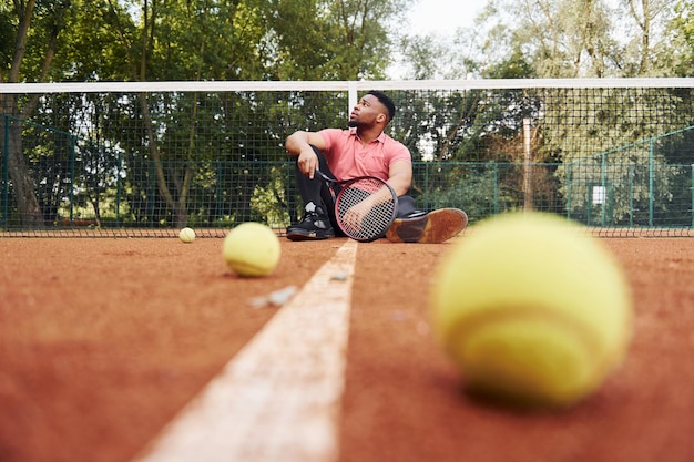 Si siede vicino alla rete e si prende una pausa L'uomo afroamericano in camicia rosa si siede con la racchetta da tennis sul campo all'aperto