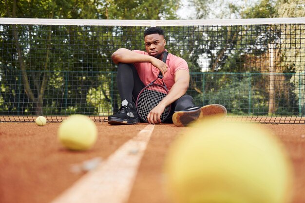 Si siede vicino alla rete e si prende una pausa L'uomo afroamericano in camicia rosa si siede con la racchetta da tennis sul campo all'aperto