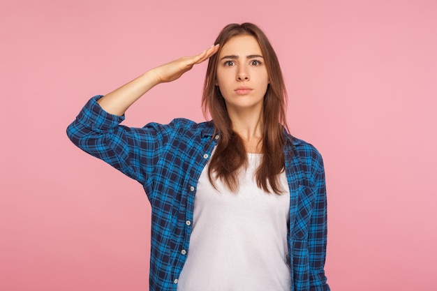 Sì, signore Ritratto di giovane donna patriottica sicura di sé in camicia a scacchi che saluta il comandante con uno sguardo attento e responsabile pronto a obbedire all'ordine studio indoor girato isolato su sfondo rosa