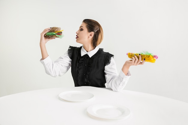 Siamo quello che mangiamo. La donna mangia hamburger e hot dog in plastica, concetto di eco. Ci sono così tanti polimeri che ne siamo fatti. Disastro ambientale, moda, bellezza, cibo. Perdere organico.