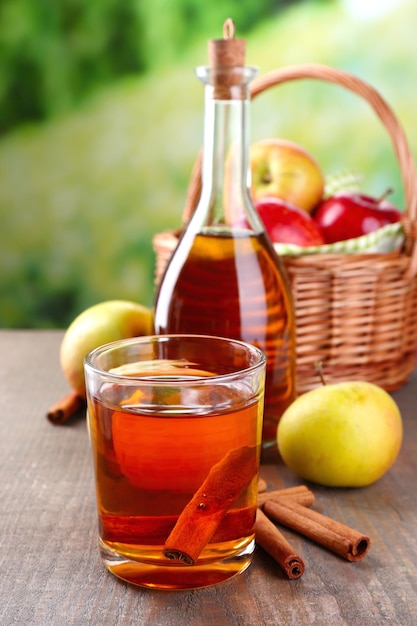 Sidro di mele in vetro e bottiglia con bastoncini di cannella e mele fresche su tavola di legno su sfondo luminoso