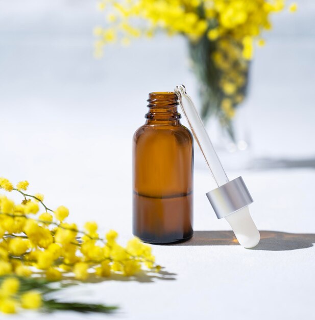 Siero viso e corpo in bottiglia su sfondo chiaro con fiori di mimosa e ombra dura primo piano Salute e bellezza