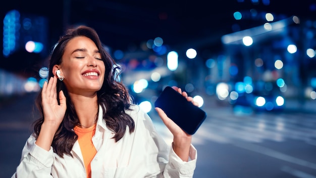 Signora allegra che ascolta la musica preferita negli auricolari tenendo lo smartphone camminando di notte sulla strada della città