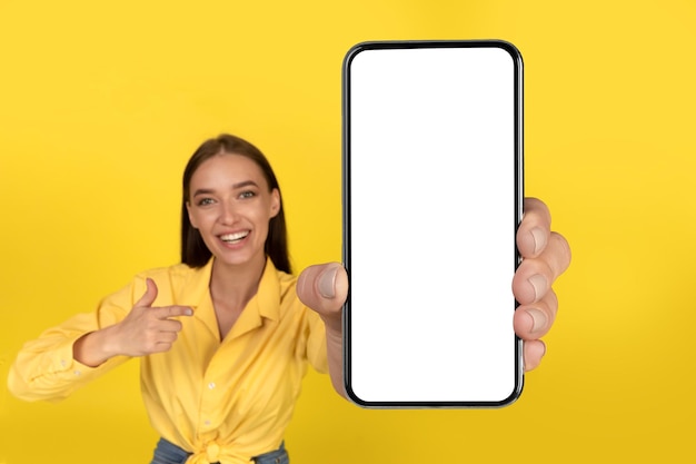 Signora allegra che mostra cellulare con schermo vuoto su sfondo giallo