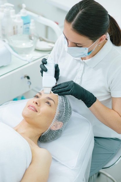 Signora allegra che sorride mentre un cosmetologo professionista esegue una pulizia del viso ad ultrasuoni
