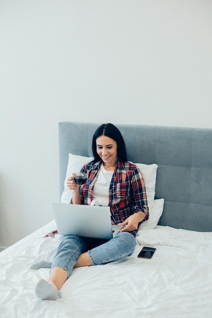 Signora allegra in abiti casual seduta sul letto con una tazza di caffè e sorridente mentre utilizza un computer portatile moderno