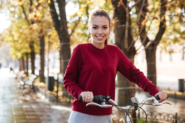 Signora allegra vestita di maglione che cammina con la sua bicicletta all'aperto