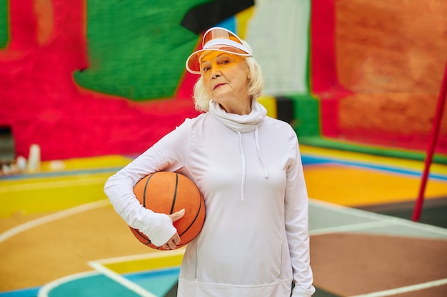 Signora anziana, sana e allegra con la palla da basket su una piazza colorata all'aperto