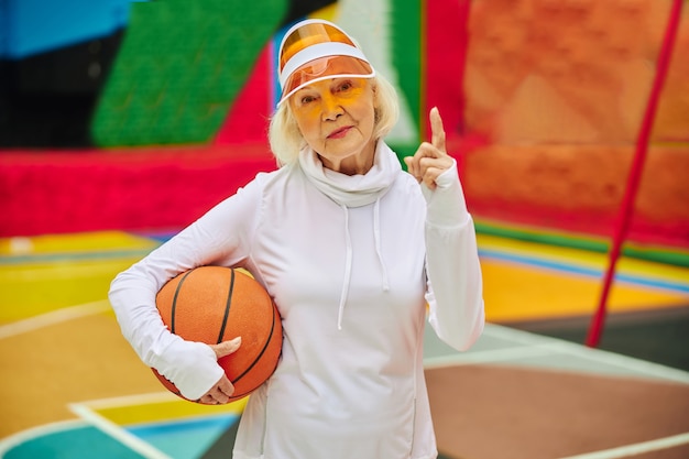 Signora anziana, sana e allegra con la palla da basket su una piazza colorata all'aperto