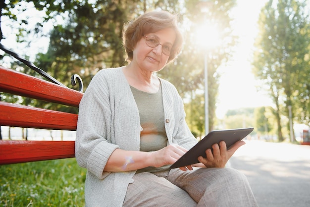 Signora anziana sicura e positiva che posa con il tablet nel parco Donna anziana dai capelli grigi in seduta casual su una panchina del parco e che utilizza il tablet Concetto di connessione wireless