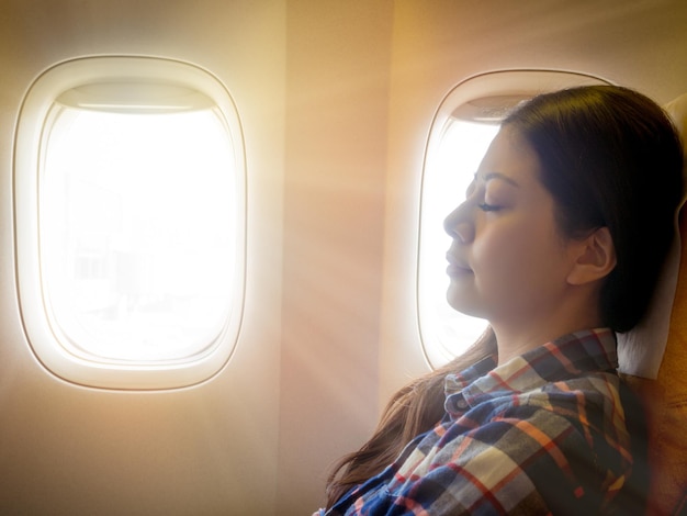 signora asiatica casual bruna stanca che fa un pisolino sul comodo sedile della cabina mentre vola viaggiando con il sole dal cielo esterno.