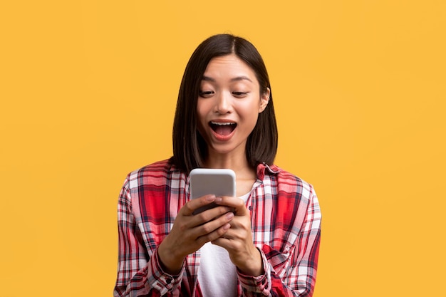 Signora asiatica sorpresa che utilizza smartphone guardando lo schermo con la bocca aperta che legge grandi notizie su sfondo giallo